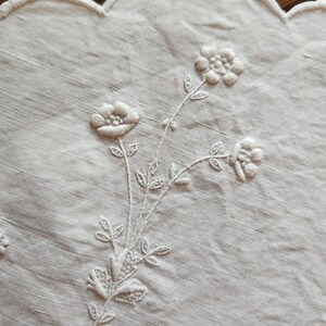 Op de afbeelding: Een gebroken wit linnen textiel met delicate toon-op-toon bloemenborduursels. De stof heeft een geschulpte rand en toont een centraal botanisch motief met drie kleine bloemen, stengels en bladeren gestikt met bijpassende draad.