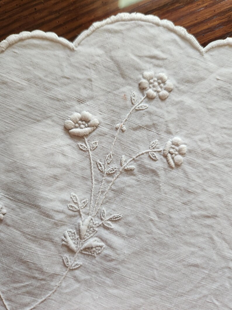 Op de afbeelding: Een gebroken wit linnen textiel met delicate toon-op-toon bloemenborduursels. De stof heeft een geschulpte rand en toont een centraal botanisch motief met drie kleine bloemen, stengels en bladeren gestikt met bijpassende draad.