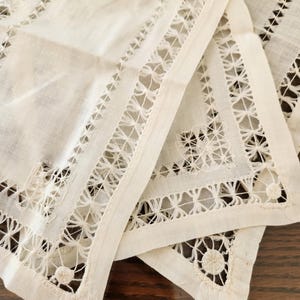 May include: Close-up of several white linen napkins with intricate cutwork and embroidered edges. The napkins are folded, revealing detailed geometric and floral patterns. The fabric appears to be slightly off-white, with a soft texture.