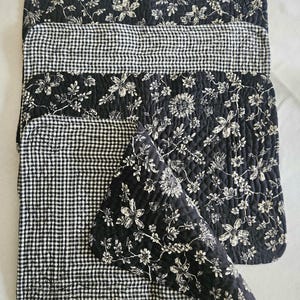 May include: A set of quilted placemats and a napkin. The placemats feature a black and white floral pattern and a black and white gingham pattern. The napkin has the floral pattern. The placemats and napkin are made of a soft, quilted fabric.