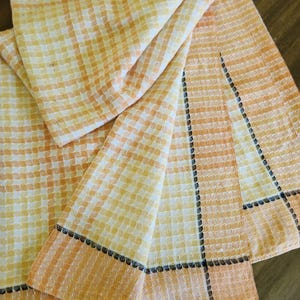 May include: A waffle-weave kitchen towel featuring a light orange and white checkered pattern. The towel has a wide orange border along the edges, accented by a decorative black stitched trim that creates a distinct rectangular frame design.