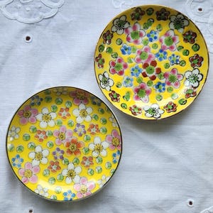May include: Two small, round, yellow plates with floral designs. The plates have a gold trim and are decorated with colorful flowers in shades of pink, white, blue, and red. The plates are on a white, textured surface.