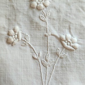 Op de afbeelding: Een stuk gebroken wit textiel met witte bloemenborduursels. Het ontwerp bestaat uit drie afzonderlijke bloemen met verhoogde, opgevulde bloemblaadjes en stengels met kleine, gedetailleerde blaadjes. De stof heeft een zichtbare, licht geweven structuur.