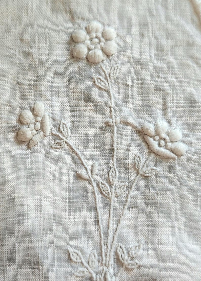 Op de afbeelding: Een stuk gebroken wit textiel met witte bloemenborduursels. Het ontwerp bestaat uit drie afzonderlijke bloemen met verhoogde, opgevulde bloemblaadjes en stengels met kleine, gedetailleerde blaadjes. De stof heeft een zichtbare, licht geweven structuur.