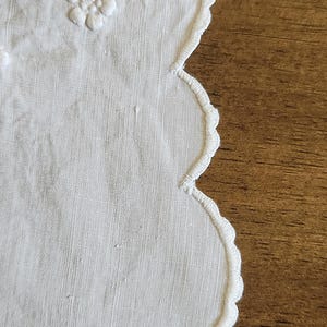 Op de afbeelding: Een wit stoffen kleedje met een geschulpte rand en geborduurde stiksels. Het textieloppervlak toont delicaat wit bloemenborduurwerk met verhoogde bloemblaadjes en kleine stippen, met een fijn weefsel en een strak afgewerkte rand.