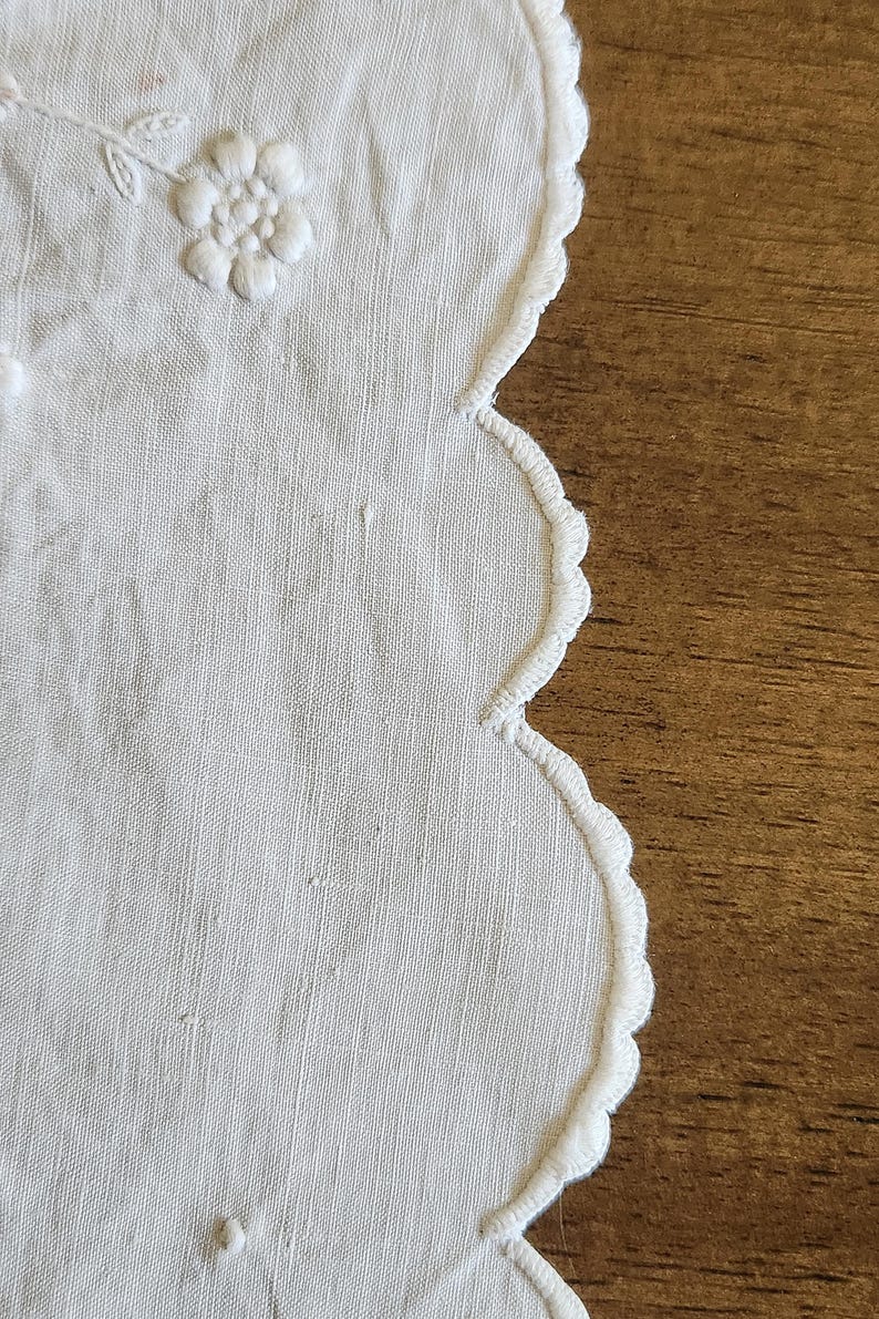 Op de afbeelding: Een wit stoffen kleedje met een geschulpte rand en geborduurde stiksels. Het textieloppervlak toont delicaat wit bloemenborduurwerk met verhoogde bloemblaadjes en kleine stippen, met een fijn weefsel en een strak afgewerkte rand.