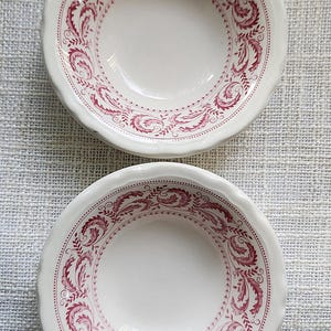 May include: Two white ceramic bowls with a scalloped edge and a burgundy floral design. The bowls have a detailed pattern around the rim and inside the bowl. The bowls are on a light gray textured surface.
