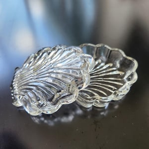 May include: A clear glass trinket dish shaped like a scallop shell, consisting of two matching pieces. The glass features a molded, ribbed texture radiating from the hinge area to the scalloped edges, creating a translucent and reflective surface.