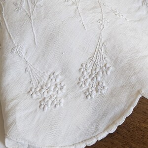 Op de afbeelding: Een wit textiel met delicate toon-op-toon bloemenborduursels. De stof heeft een geschulpte rand met ingewikkeld stikwerk. Het ontwerp bestaat uit slanke stelen die leiden naar trossen kleine, gedetailleerde bloesems in wit garen.