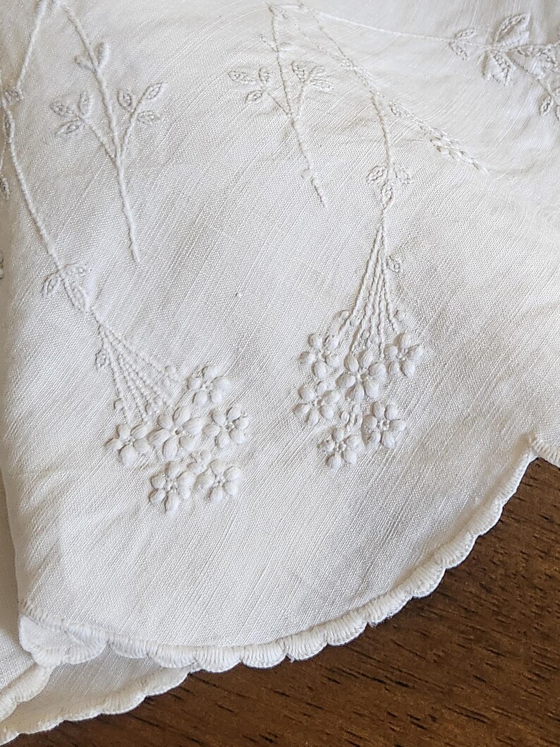 Op de afbeelding: Een wit textiel met delicate toon-op-toon bloemenborduursels. De stof heeft een geschulpte rand met ingewikkeld stikwerk. Het ontwerp bestaat uit slanke stelen die leiden naar trossen kleine, gedetailleerde bloesems in wit garen.