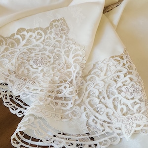 May include: Ivory-colored tablecloth with intricate lace detailing. The lace features floral and geometric patterns along the edges and corners. The fabric has a subtle sheen, and the design is visible in a close-up shot.