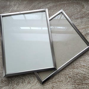 May include: Two rectangular silver picture frames with glass fronts. One frame has a white backing, the other a beige backing. The frames are slightly overlapping, with a textured surface in the background.