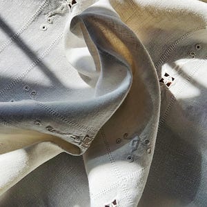 May include: Close-up of a cream-colored, intricately embroidered fabric. The fabric features geometric patterns, cut-out designs, and delicate stitching. The folds and shadows create a sense of depth and texture, highlighting the craftsmanship.