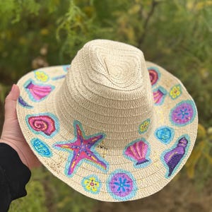 Puede incluir: Un sombrero de paja de ala ancha decorado con conchas marinas pintadas, estrellas de mar y otros diseños de temática oceánica en rosa, morado, azul y amarillo.