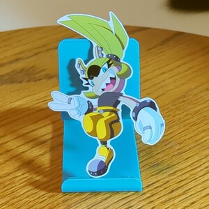 May include: A teal blue phone stand with a cartoon character cutout. The character is a yellow and brown anthropomorphic animal with a black eye patch and a toothy grin. The character is wearing yellow pants and white gloves.
