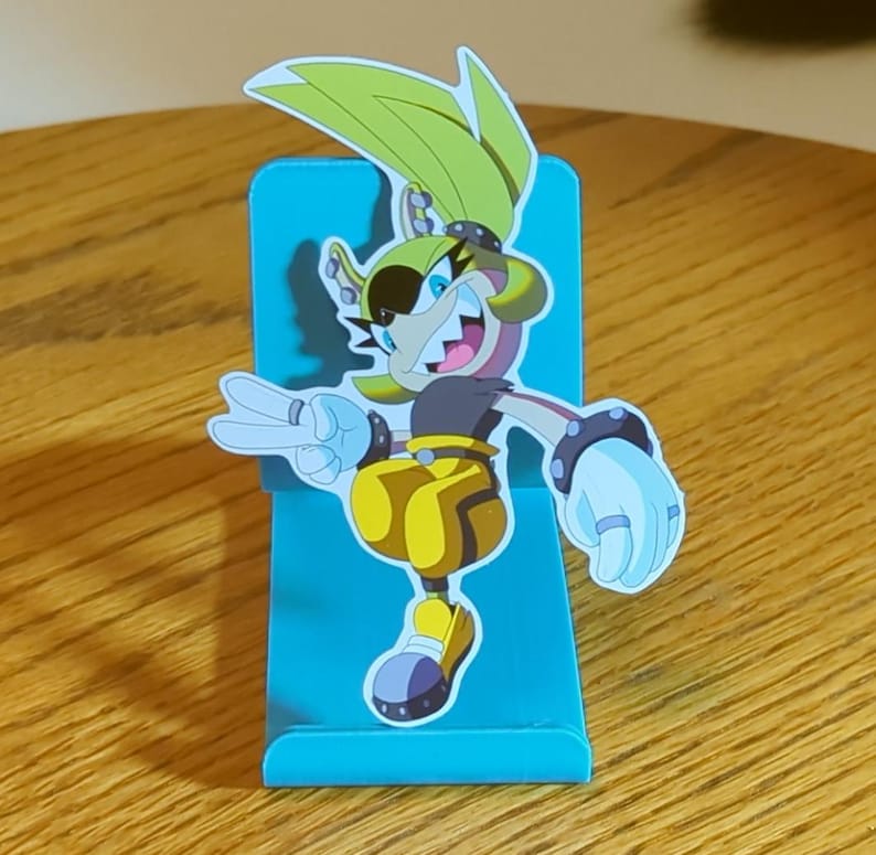 May include: A teal blue phone stand with a cartoon character cutout. The character is a yellow and brown anthropomorphic animal with a black eye patch and a toothy grin. The character is wearing yellow pants and white gloves.