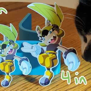 May include: Two cartoon character stickers, one 3 inches tall and the other 4 inches tall. Both characters are yellow and green with black eyes and a black eyepatch. They are wearing black and yellow outfits and have a white glove on each hand. The characters are on a blue background.
