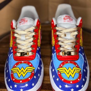 May include: Custom Nike Air Force 1 trainers with a Wonder Woman theme. The shoes are in blue, red, and yellow, featuring the Wonder Woman logo, stars, and gold laces. The trainers are a vibrant design.