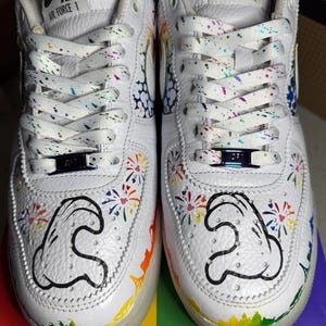 May include: White Air Force 1 sneakers with colorful accents. The shoes feature rainbow-colored details, including the laces, soles, and a design on the side. The design includes a black outline of a cartoon character.