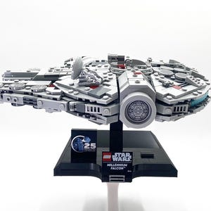 Könnte beinhalten: Ein graues LEGO Millennium Falcon Modell mit roten und blauen Details, das auf einem schwarzen Sockel mit dem LEGO Star Wars Logo und dem Text "Millennium Falcon" präsentiert wird. Der Sockel zeigt auch ein 25-jähriges Jubiläumslogo.