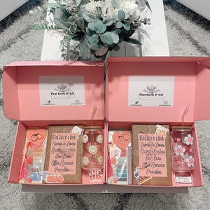 May include: Two pink gift boxes with lids open, revealing blind date book boxes. Each box contains a book, a tumbler, a bookmark, and a keychain. The book is wrapped in brown paper with a pink ribbon and a tag that says "Blind Date w/ a Book". The tumblers are clear glass with a smiley face design and a floral design. The bookmarks are pink and white with a tassel. The keychains are heart-shaped with a pink and white design. The boxes are on a white surface with a bouquet of white flowers in the background.