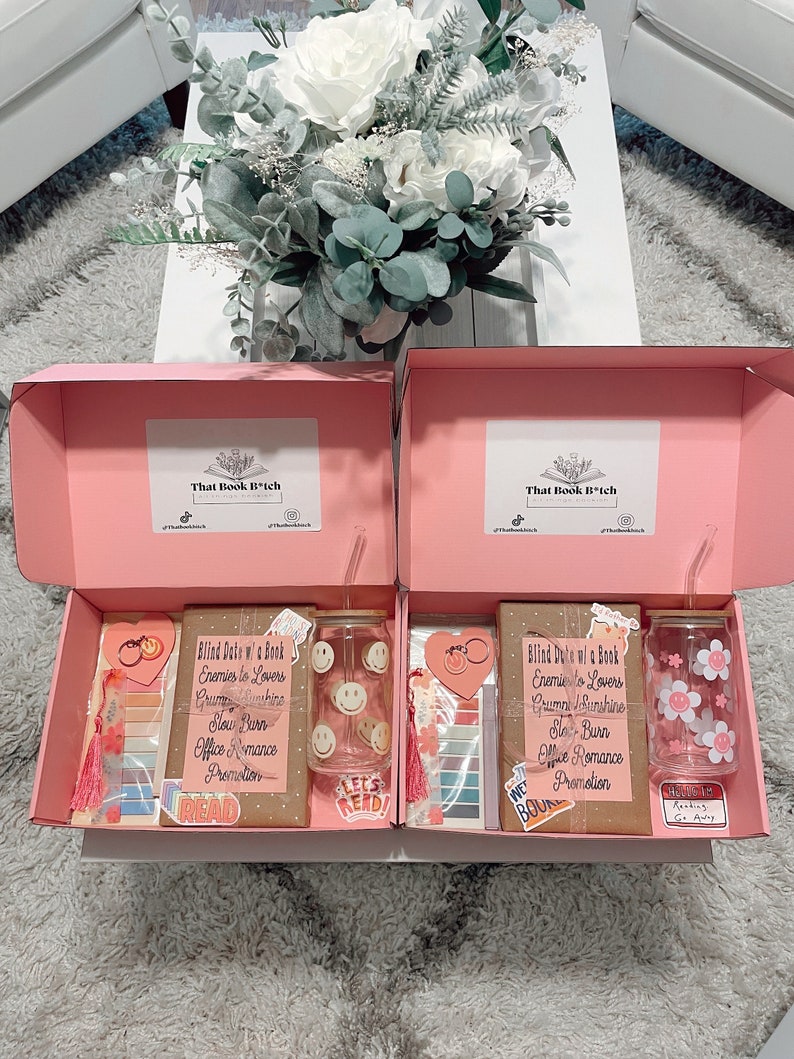 May include: Two pink gift boxes with lids open, revealing blind date book boxes. Each box contains a book, a tumbler, a bookmark, and a keychain. The book is wrapped in brown paper with a pink ribbon and a tag that says "Blind Date w/ a Book". The tumblers are clear glass with a smiley face design and a floral design. The bookmarks are pink and white with a tassel. The keychains are heart-shaped with a pink and white design. The boxes are on a white surface with a bouquet of white flowers in the background.
