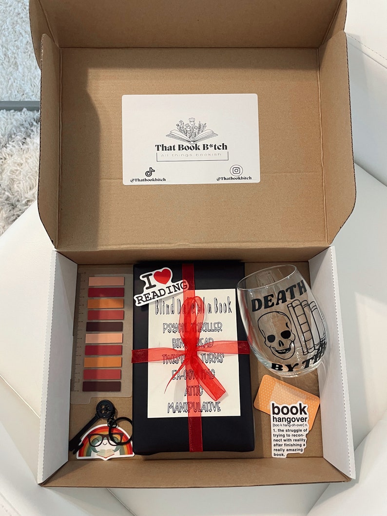 May include: A brown cardboard box with a black book wrapped in red ribbon, a wine glass with a skull and crossbones design, a sticker that says "I love reading", a sticker that says "Blind Date with a Book", a sticker that says "Death by Book", and a sticker that says "Book Hangover".