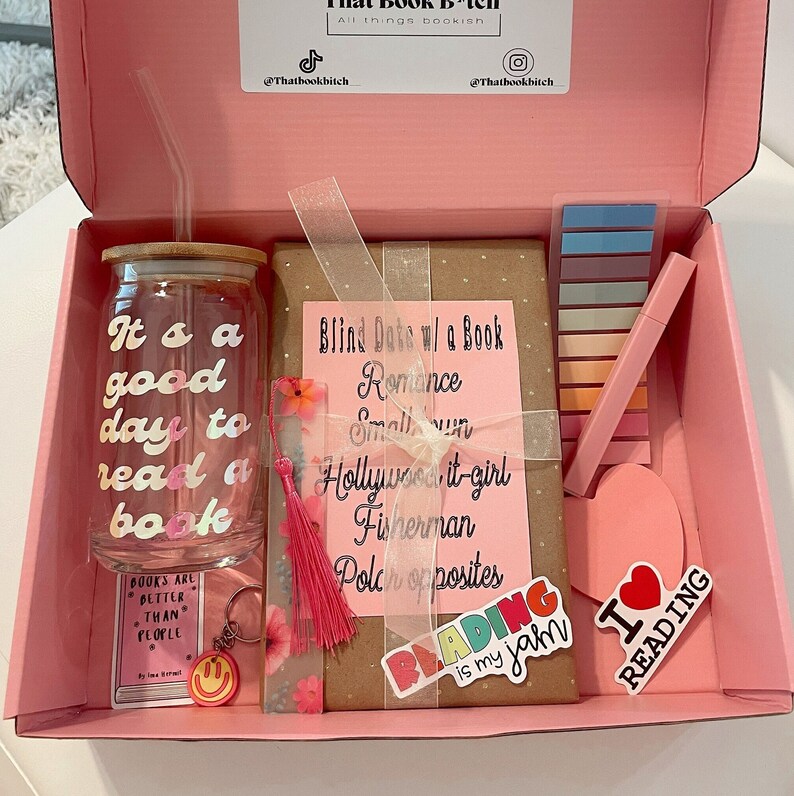 May include: A pink gift box filled with bookish items, including a glass tumbler with the text "It's a good day to read a book", a brown wrapped book with the text "Blind Date w/ a Book", a pink tassel bookmark, a set of colorful sticky notes, a pink heart-shaped sticker, and a sticker that says "I love reading".
