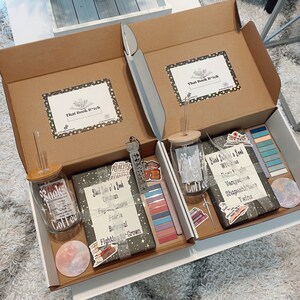 May include: Two open cardboard boxes with white interiors, each containing a bookish themed gift box. The boxes include a glass jar with a straw, a book, and various stickers and bookmarks. The boxes are labeled "That Book B*tch".