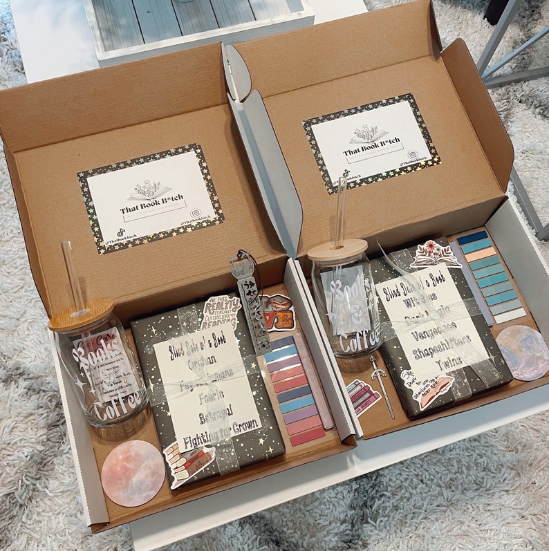 May include: Two open cardboard boxes with white interiors, each containing a bookish themed gift box. The boxes include a glass jar with a straw, a book, and various stickers and bookmarks. The boxes are labeled "That Book B*tch".