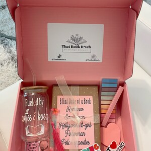 May include: A pink gift box filled with book-themed items, including a book, a tumbler with the text "Fueled by coffee & books", a bookmark, a set of colorful sticky notes, and a heart-shaped sticker that says "I love reading".
