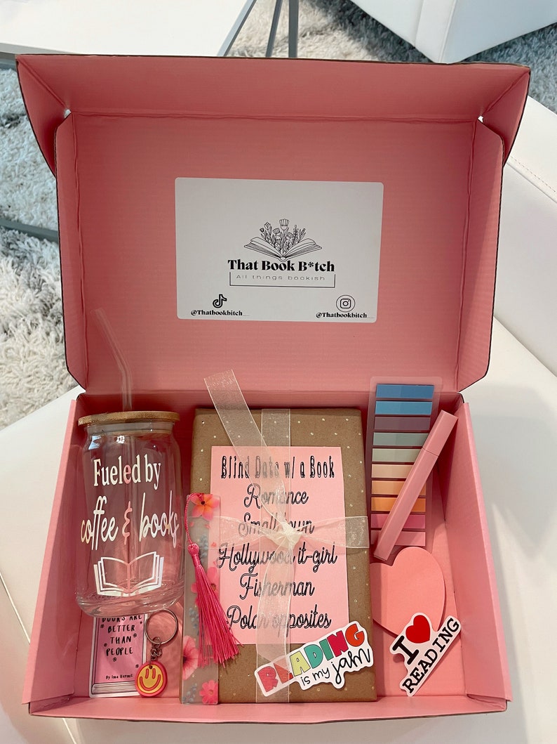 May include: A pink gift box filled with book-themed items, including a book, a tumbler with the text "Fueled by coffee & books", a bookmark, a set of colorful sticky notes, and a heart-shaped sticker that says "I love reading".