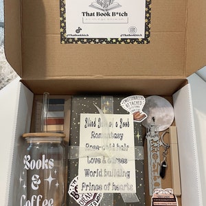 May include: A brown cardboard box with a lid open, revealing a gift box set for book lovers. The set includes a glass jar with a wooden lid that says "Books & Coffee", a book wrapped in gray paper with gold stars and the text "Blind Date w/ a Book", a bookmark, a keychain, a sticker, and a ruler.