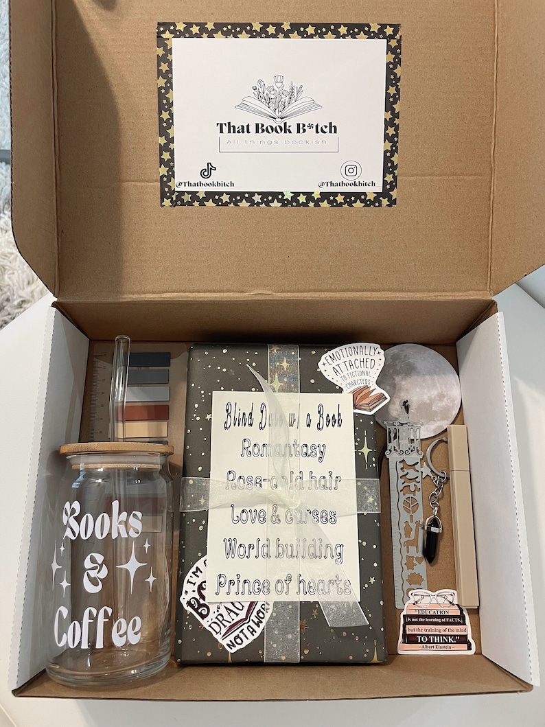 May include: A brown cardboard box with a lid open, revealing a gift box set for book lovers. The set includes a glass jar with a wooden lid that says "Books & Coffee", a book wrapped in gray paper with gold stars and the text "Blind Date w/ a Book", a bookmark, a keychain, a sticker, and a ruler.
