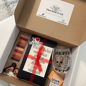 May include: A gift box filled with book-themed items, including a black book with a red ribbon, a wine glass with a skull and crossbones design, and a sticker that says "book hangover".