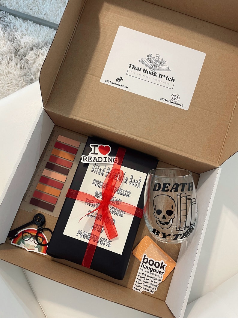 May include: A gift box filled with book-themed items, including a black book with a red ribbon, a wine glass with a skull and crossbones design, and a sticker that says "book hangover".