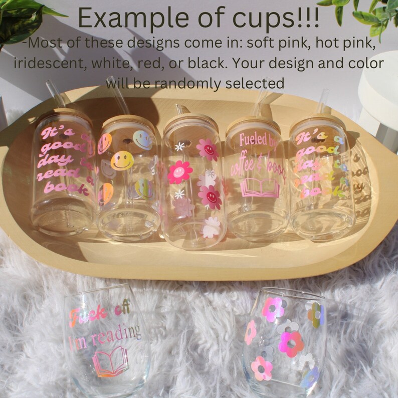 May include: A collection of seven glass cups with various designs and colors. The cups have a straw and a wooden lid. The designs include flowers, smiley faces, and text such as "It's a good day to read a book" and "Fueled by coffee & books".