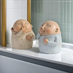 May include: Two ceramic figurines, each depicting a bald figure covering their mouth with a hand. One is in a cream-colored sweater with star details, the other in a light blue sweater. The figures have rosy cheeks and are set against a tiled background.