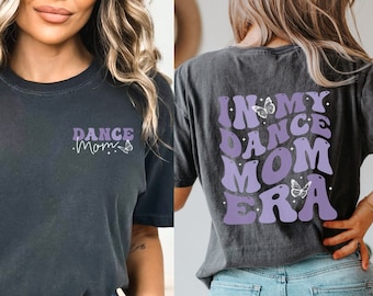 May include: Two women wearing gray t-shirts with purple text and butterfly graphics. The text on the first shirt says "Dance Mom" and the text on the second shirt says "In My Dance Mom Era".