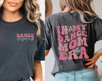May include: A dark gray t-shirt with the text "Dance Mom" in pink and white lettering with a butterfly graphic. The back of the shirt has the text "In My Dance Mom Era" in pink and white lettering with a butterfly graphic.