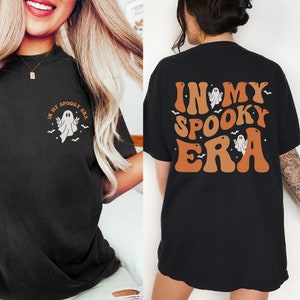May include: Black t-shirts with Halloween-themed graphics. One shirt features a front design with a ghost and the text "IN MY SPOOKY ERA". The other shirt has the same text and ghost design on the back in orange and brown.