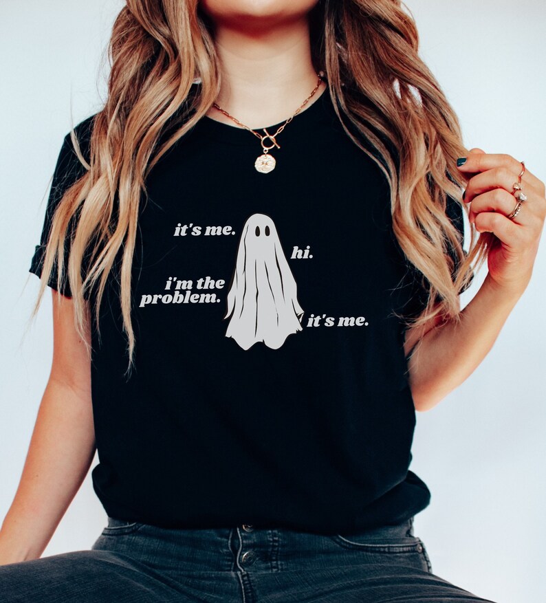 May include: A black short-sleeve t-shirt featuring a white graphic of a ghost in the center. The text printed in white reads: it's me. hi. i'm the problem. it's me. The shirt has a crew neckline and a standard fit, showcasing a simple and clean design.