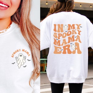 May include: A white sweatshirt featuring a small graphic on the front left chest of a ghost making a peace sign with the text "SPOOKY MAMA" above it. The back displays a large graphic with the text "IN MY SPOOKY MAMA ERA" in orange.