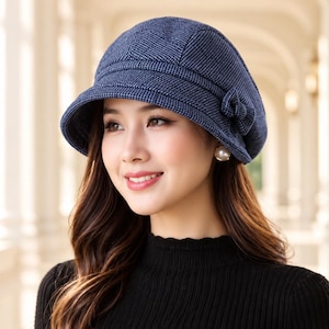 Puede incluir: Un sombrero tipo cloche de color azul oscuro con un sutil patrón de cuadrícula blanca, una copa redondeada, un ala inclinada hacia abajo y un pequeño detalle de lazo de tela decorativo en el lateral. El sombrero tiene costuras visibles y un acabado texturizado.