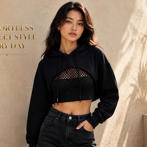 Puede incluir: Una sudadera negra corta con capucha, mangas largas y cordones, sobre un top corto de rejilla negro. El texto "EFFORTLESS STREET STYLE EVERY DAY" está impreso en dorado en el lado izquierdo. El conjunto se completa con unos vaqueros negros.