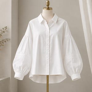 May include: A crisp white button-down shirt featuring a classic collar, a front button placket, and voluminous long sleeves with gathered cuffs. The shirt has a high-low hemline and horizontal pleat detailing on the lower portion of the sleeves.