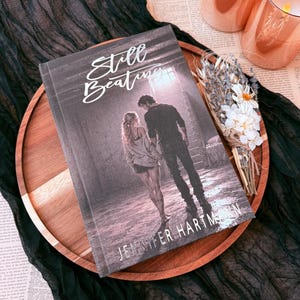 May include: A hardcover book titled Still Beating by Jennifer Hartmann featuring a cover illustration of a person with long hair and a person with short hair standing together in a dimly lit room, positioned on a round wooden tray with dried flowers.
