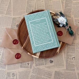 K&ouml;nnte beinhalten: Ein t&uuml;rkisfarbenes Buch mit wei&szlig;em Text und einem dekorativen Umschlagdesign liegt auf einem Holztablett, zusammen mit zwei versiegelten Umschl&auml;gen und einem kleinen Strau&szlig;. Die Umschl&auml;ge sind mit rotem Wachs versiegelt.