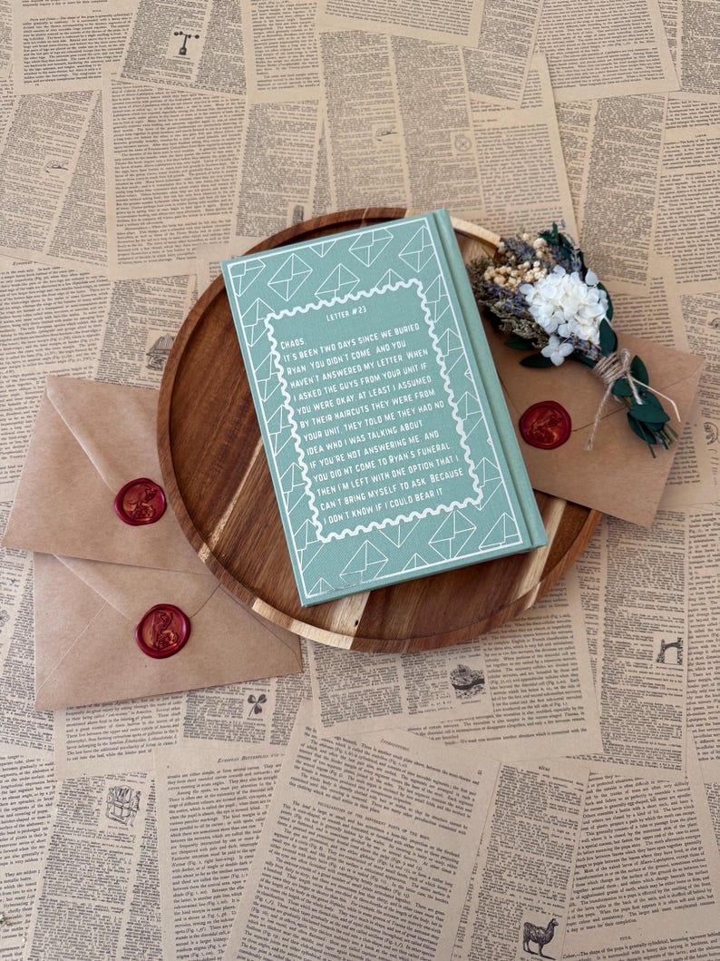 K&ouml;nnte beinhalten: Ein t&uuml;rkisfarbenes Buch mit wei&szlig;em Text und einem dekorativen Umschlagdesign liegt auf einem Holztablett, zusammen mit zwei versiegelten Umschl&auml;gen und einem kleinen Strau&szlig;. Die Umschl&auml;ge sind mit rotem Wachs versiegelt.