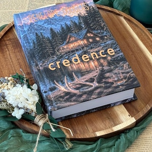 May include: A hardcover book titled "Credence" by Penelope Douglas. The cover features a mountain landscape with a cabin, lake, and antlers. The book is on a wooden tray with a bouquet of dried flowers.